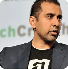 Man with short dark hair wearing a dark blazer and black shirt speaking against a blurred background.